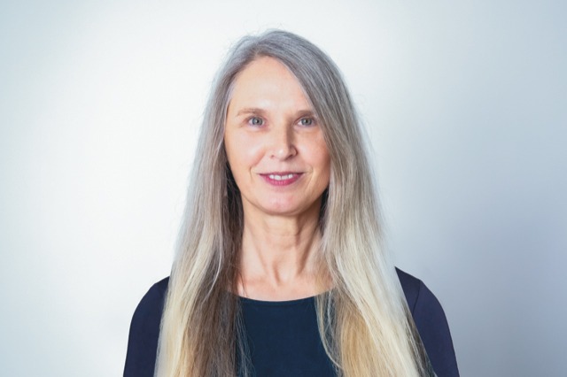 Martina Berger. Frau mit langen, grau-blonden Haar, in einem unken Oberteil.